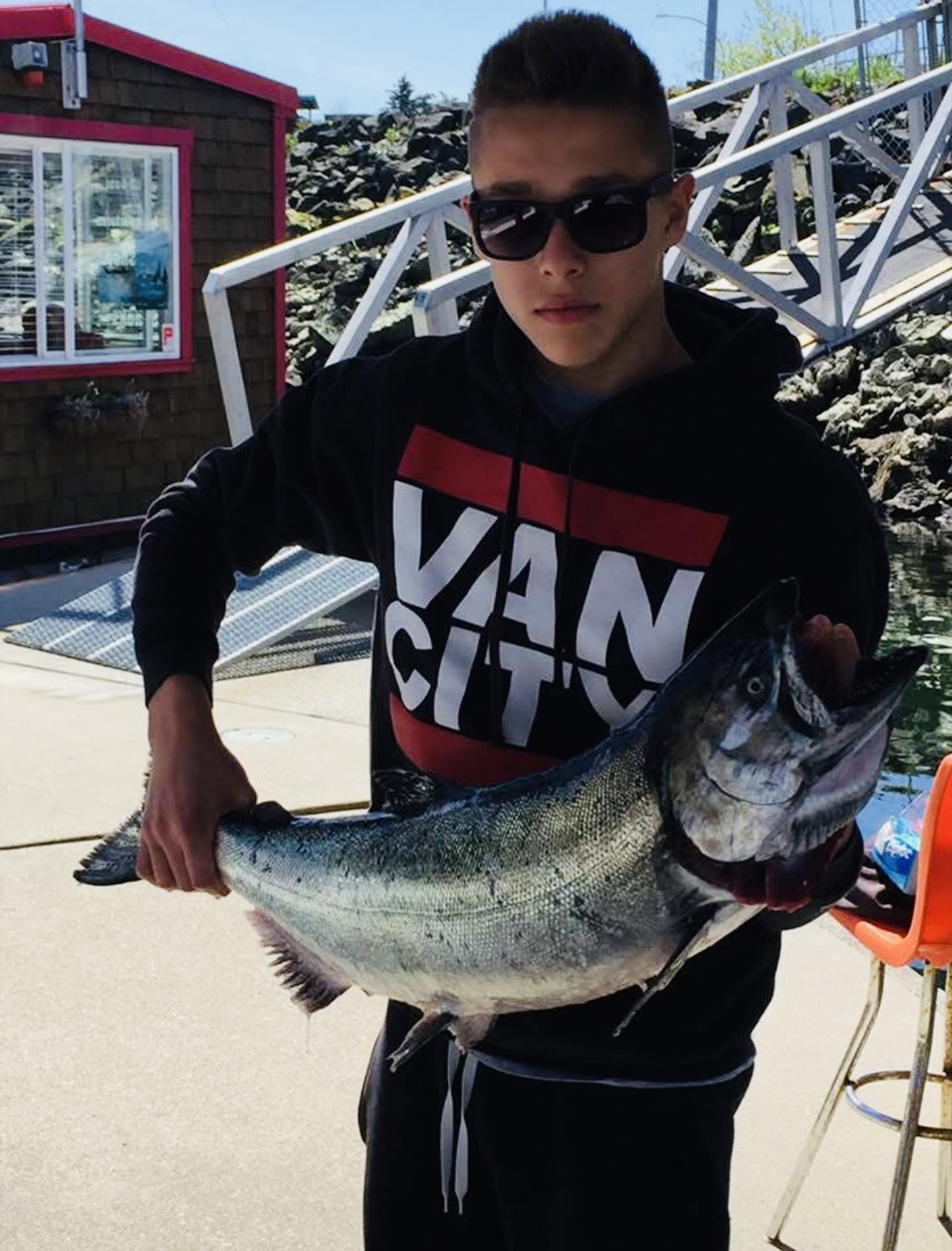 A young man holding a fish on a dock, posing for the camera. Behind him is a small building, a ramp to the dock, and a rocky embankment.